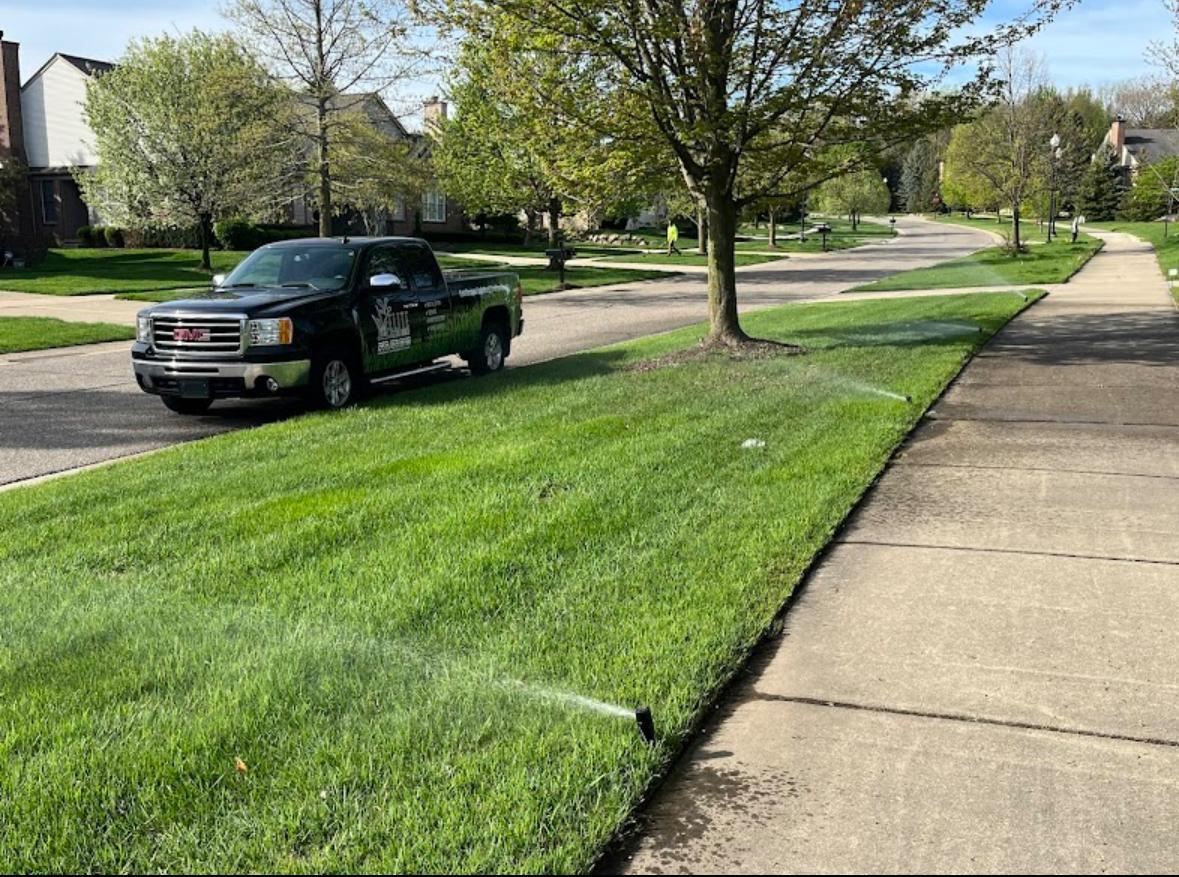 Elite Sprinkler Systems truck on a Metro Detroit residential street with active sprinklers running across a lush green lawn.